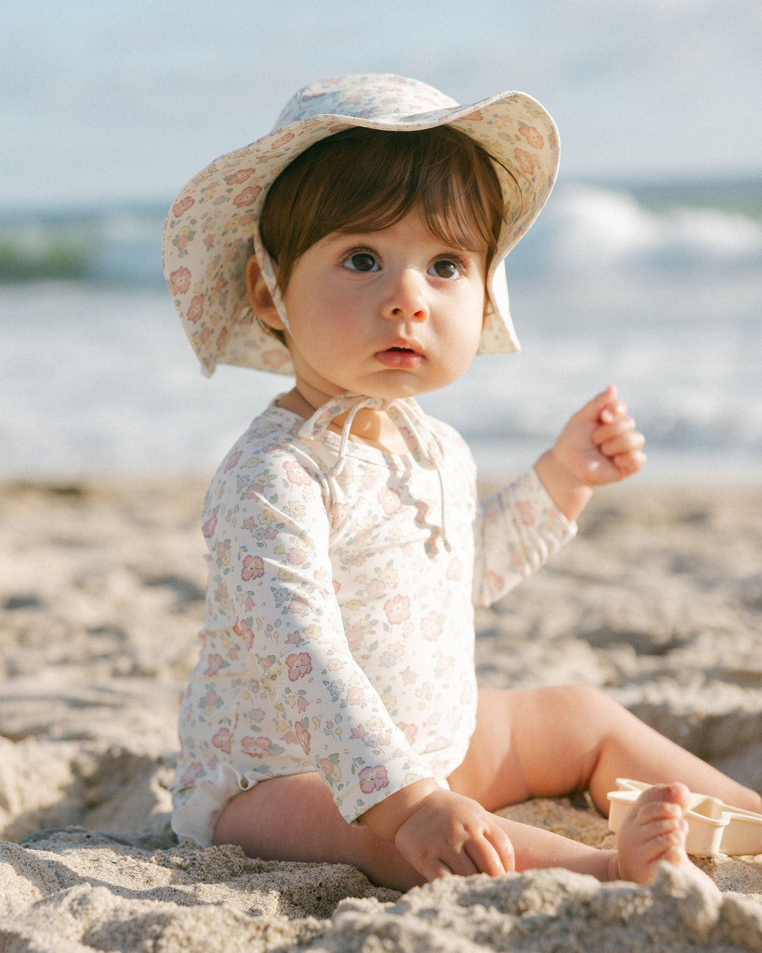 baby modeling the pastel floral rashguard and matching hat. the baby is looking up and you can see the print in day light. 