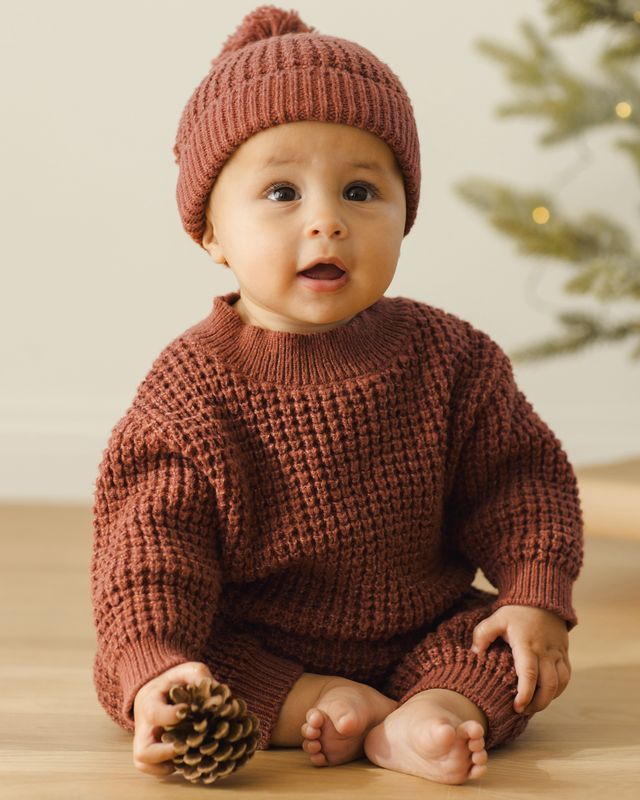 Baby wearing a cranberry knitted sweater and hat, sitting on a wooden floor with a blurred background.