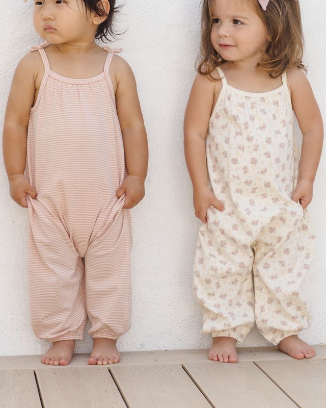 Two children wearing pink and floral-patterned rompers against a white wall.