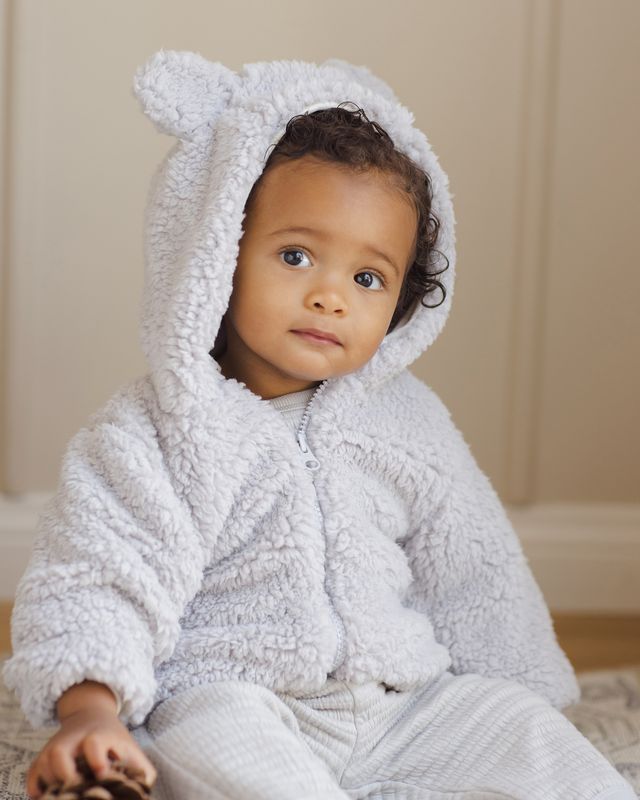 Baby wearing a light blue/grey hooded jacket with bear ears on a neutral background