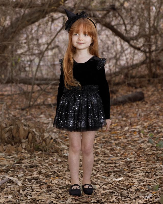 Young girl in a black dress with sequins and a witch hat standing in a forest.