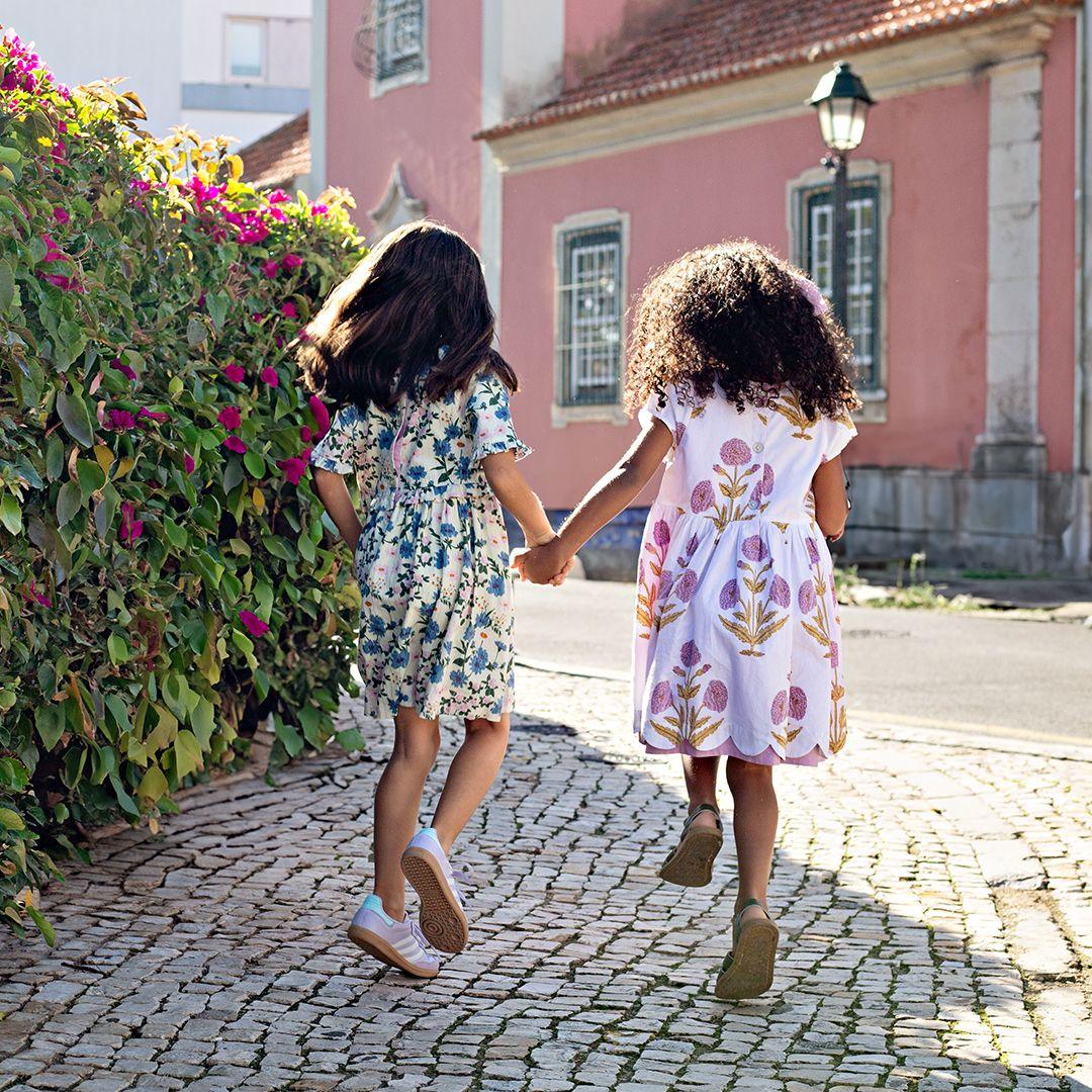 two young girls skipping down the cobblestone streets. one girl is wearing a white dress with lavender marisols and one dress with blue flowers and lavender flowers 