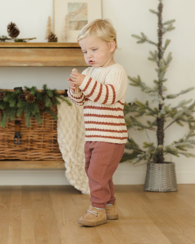 Child wearing a striped sweater and pink pants standing in a room with a Christmas tree and fireplace.