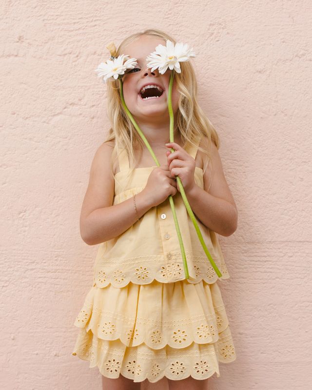 Young girl in a yellow two piece outfit with eyelet detailing holding flowers against a pink background