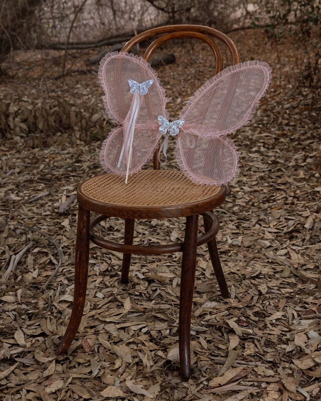 Pink fairy wings on a wooden chair in a natural setting with dry leaves and branches.