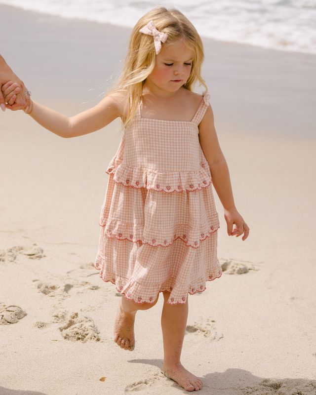 Young girl in a pink checkered dress walking on a sandy beach.