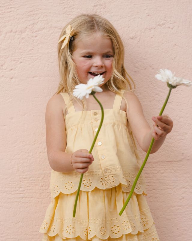 Young girl in a yellow two piece outfit with eyelet detailing holding two white flowers against a pink wall