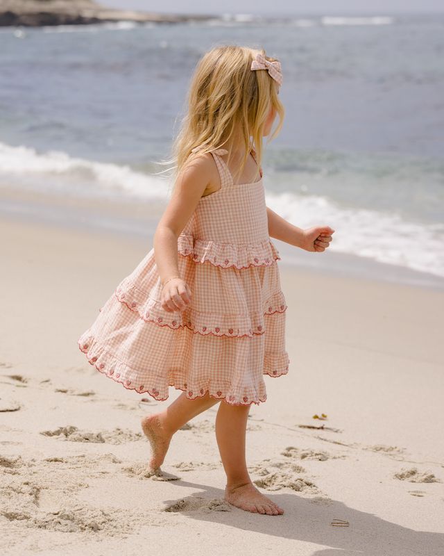 Young girl in a pink checkered dress walking on a sandy beach with ocean waves in the background.