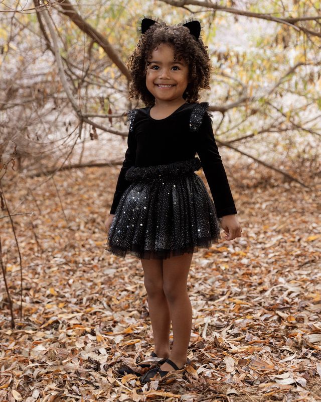 Child in a black cat costume standing in a forest with bare trees and fallen leaves.