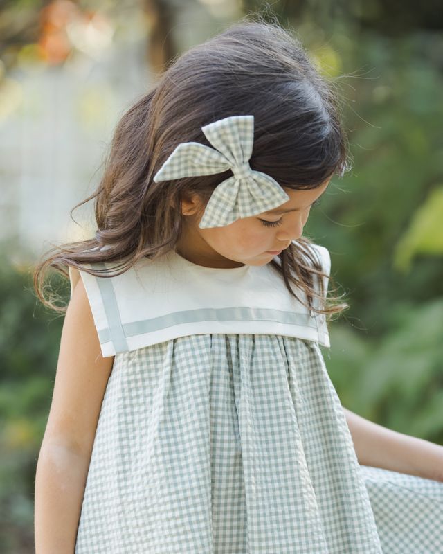 Young girl wearing a green checkered dress with a large bow in her hair, standing outdoors.