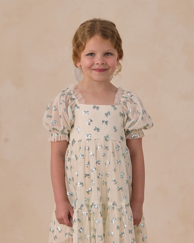 Young girl wearing a beige dress with tiny blue sequin bows against a beige background