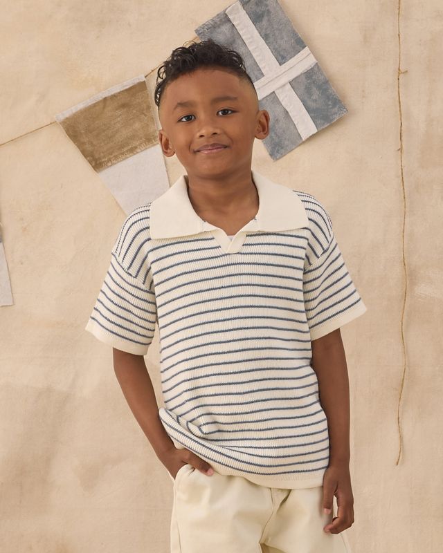 Child wearing a striped shirt and beige pants against a neutral wall with decorative elements.