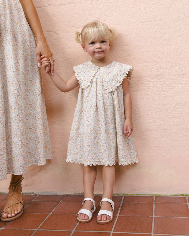 Child wearing a floral dress standing next to an adult against a pink wall.