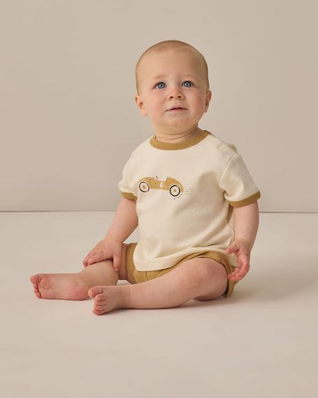 Baby sitting on a beige floor wearing a white and brown outfit with a car design.