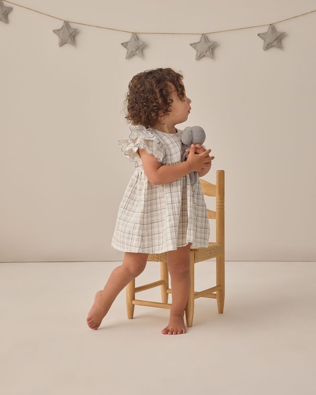 Child in a checkered dress holding a teddy bear, standing on a chair against a neutral background with star decorations.