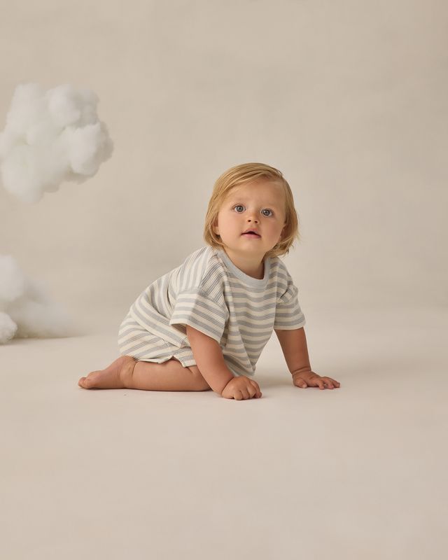 Baby in a striped outfit sitting on a beige surface with a cloud-like object in the background
