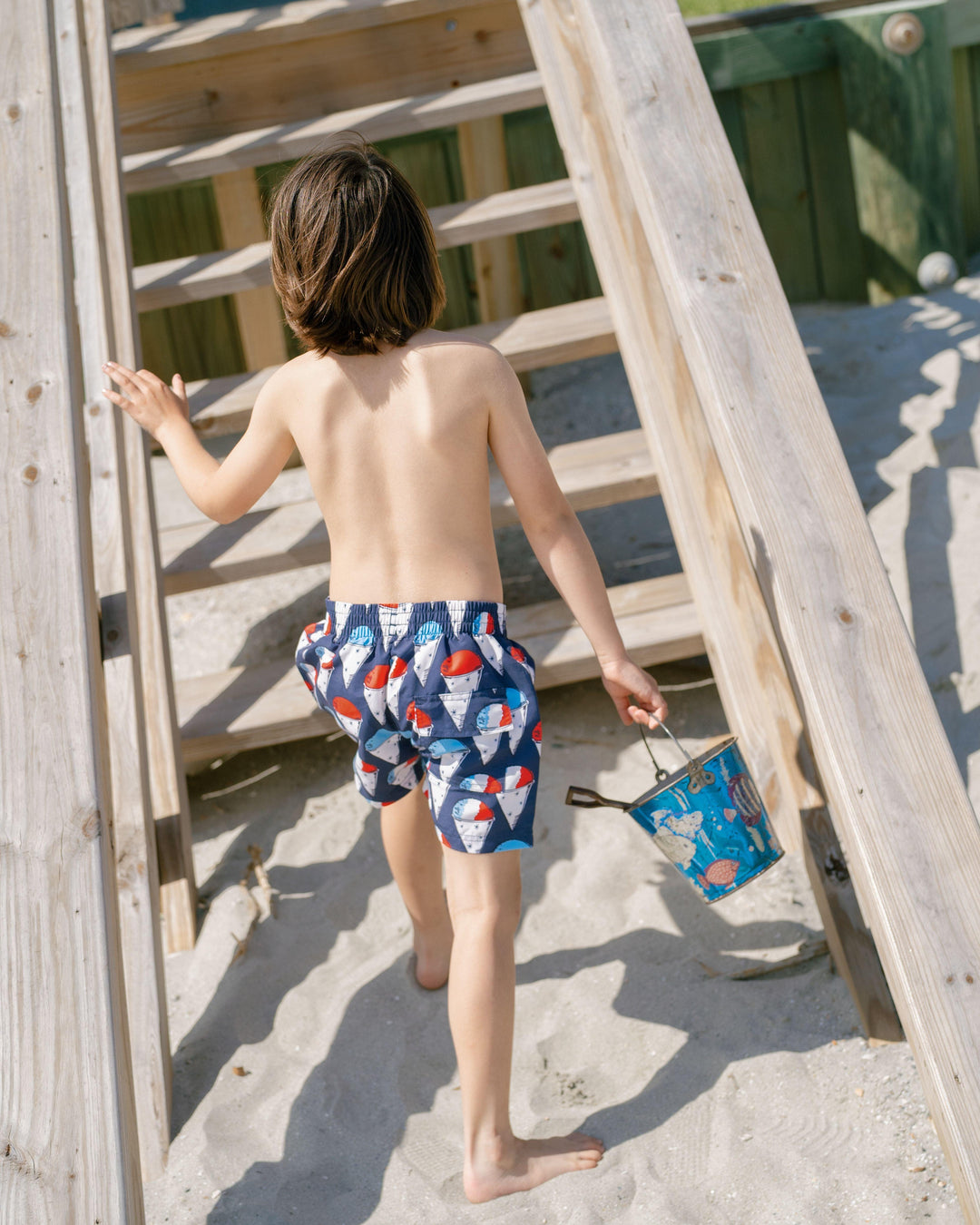 little boy wearing navy wim trunks with red white and blue snow cones printed on them. the boy is walking up the stpes and you can see the trunks from behind. 