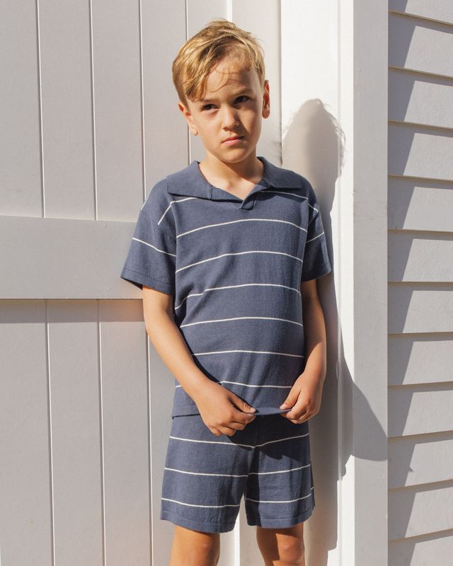 Young boy wearing a blue striped shirt and shorts against a light wooden panel background