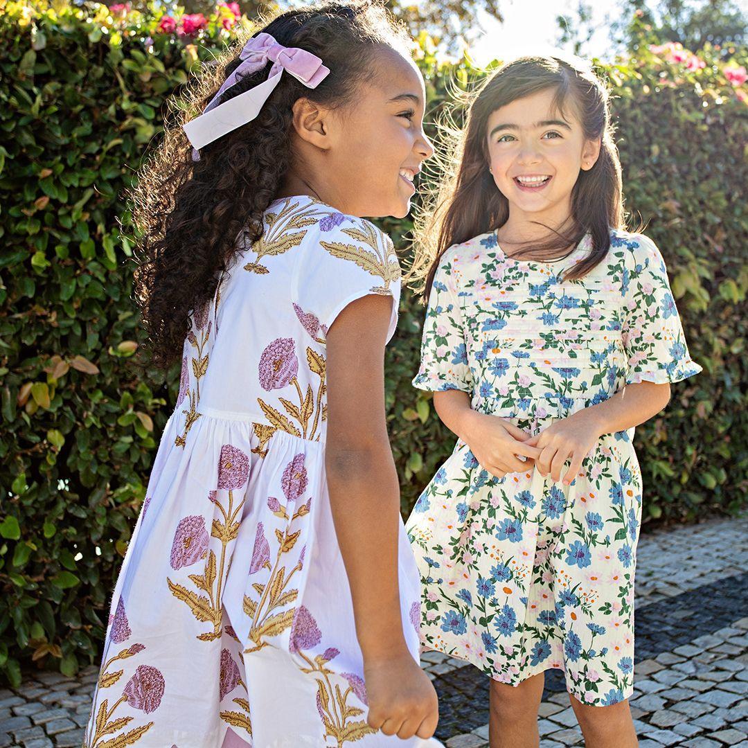 the young girls aughing and wearing their dresses in the sunlight as the sun hits them. one in white with blue flowers and one in white with lavender flowers. 