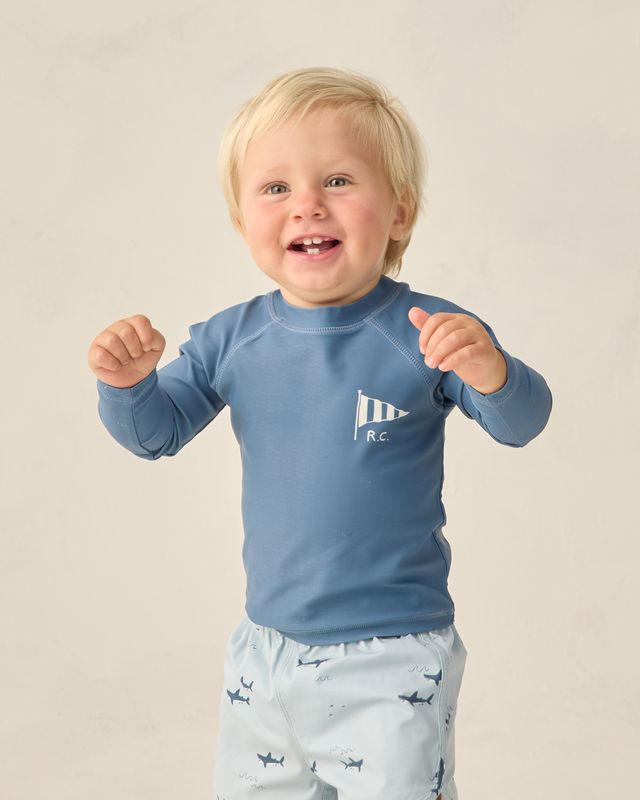 Child wearing a blue long-sleeve shirt with a logo and light-colored shorts with sharks patterns on a plain background