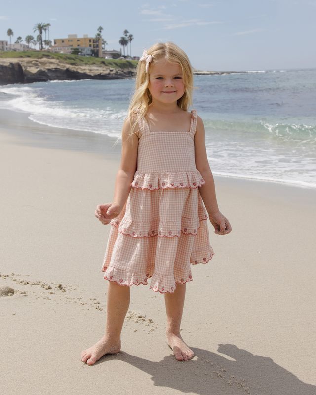 Young girl in a pink dress standing on a sandy beach with ocean waves in the background.