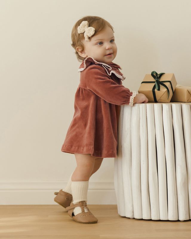 Child in a rust-colored dress standing next to a white radiator with wrapped gifts.