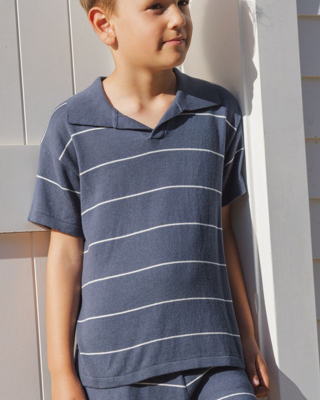 Child wearing a blue striped polo shirt standing against a light-colored wall.