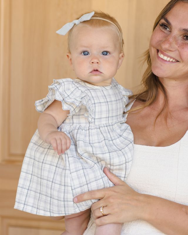 Woman holding a baby wearing a blue gray plaid dress indoors.
