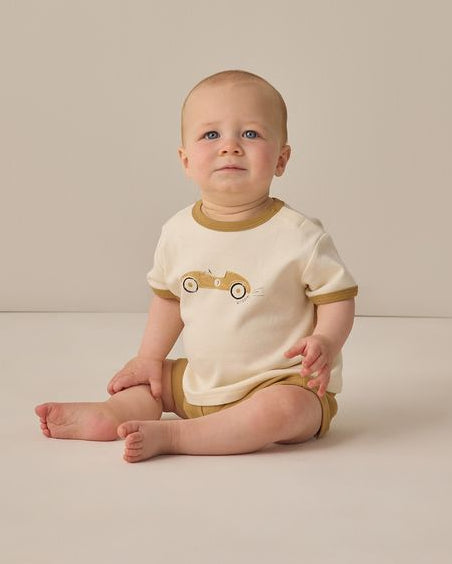 Baby wearing a white and brown outfit with a car design, sitting on a beige surface.