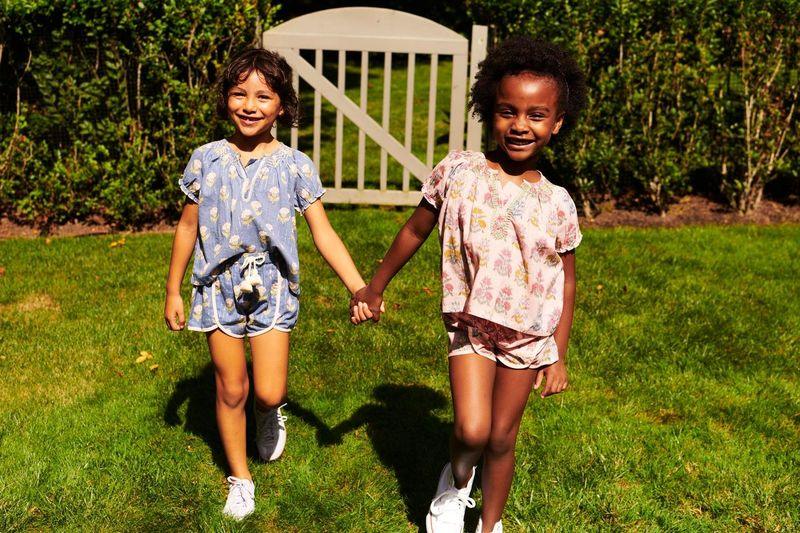 two little girls walking holding hands, both of them wearing the ava top and millie short set. one in blue and the other in pink.