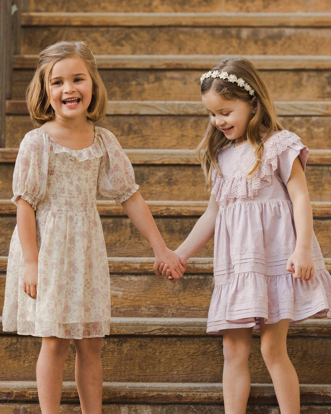 two little girls laughing and holding hands one ittel girl dressed in lavender and the other in the chiffon millie dress. 