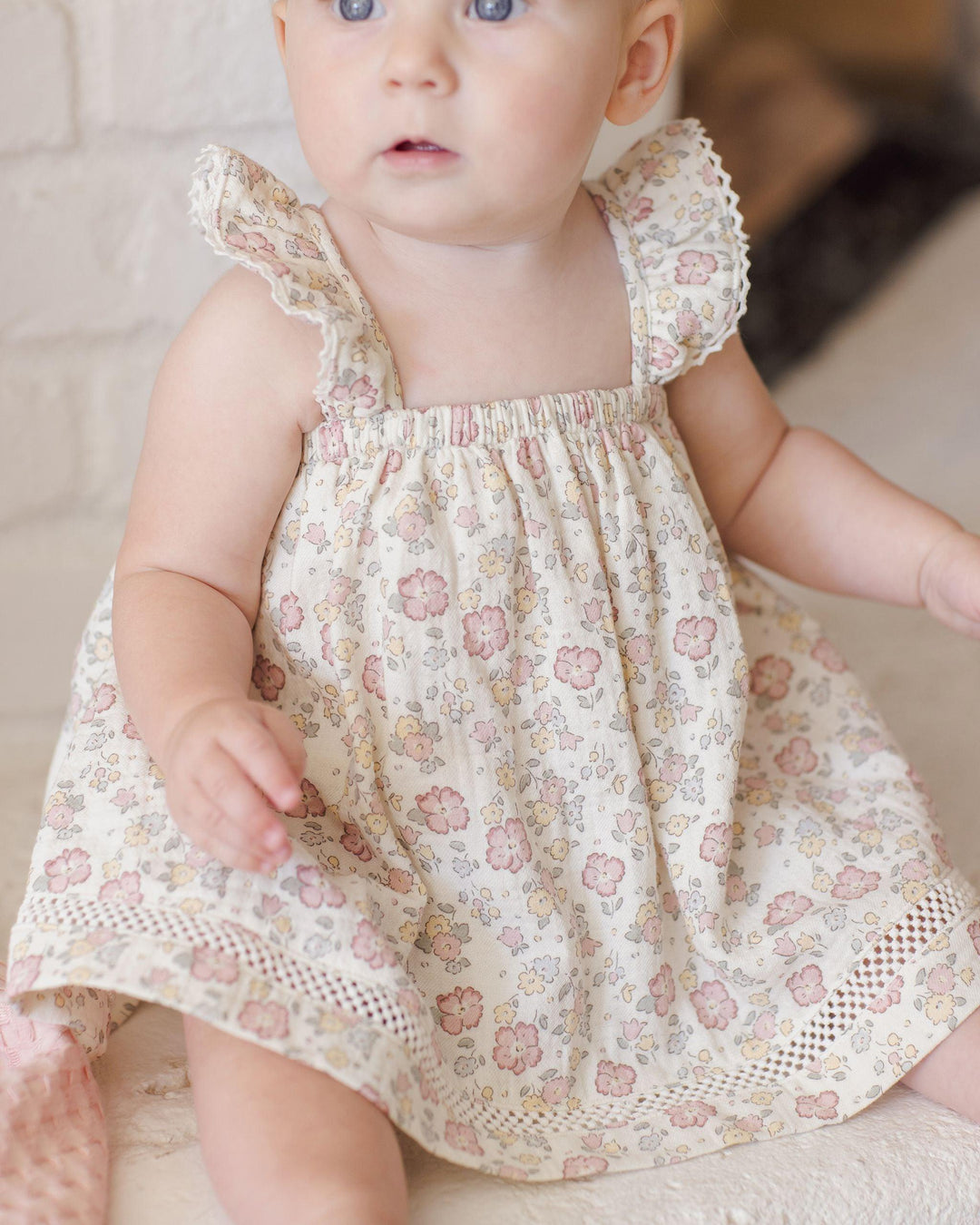 baby wearing a pastel floral dress in the ruffle tank style. lattice/lace popping through at the bottom of the dress. 