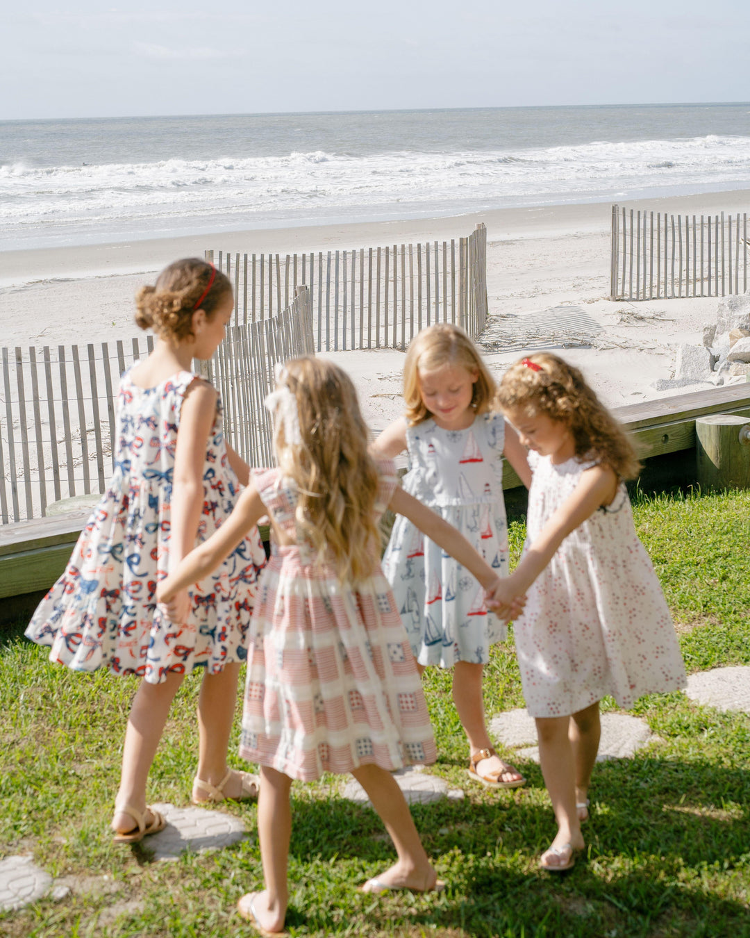 4 little girls holding hands and walking around in circles on the grass in front of the beach. 