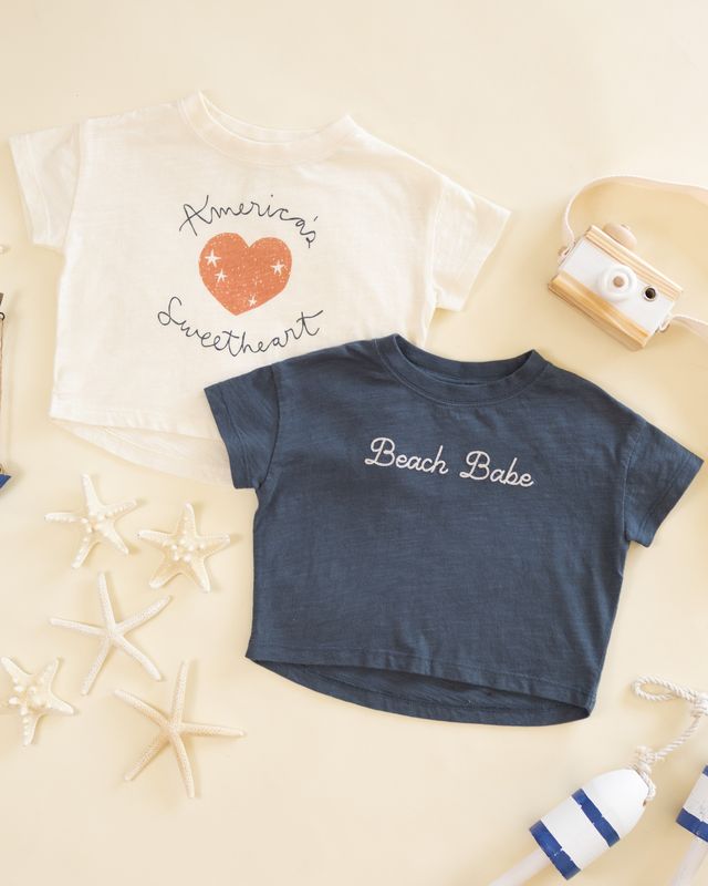 Blue t-shirt with 'Beach Babe' text on a beige background with starfish and a seashell.