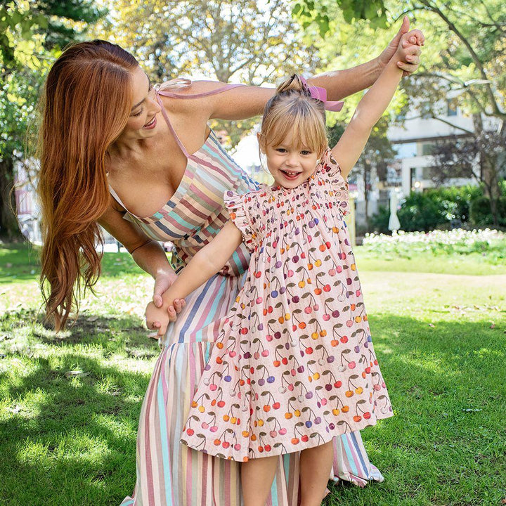 Little girl modeling pink dress with multi colored cherries all over the dress. she is playing with her mom outside in a coordinating striped dress