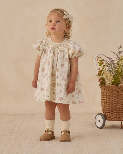 Young girl in a floral dress standing next to a wicker cart with flowers on a beige background