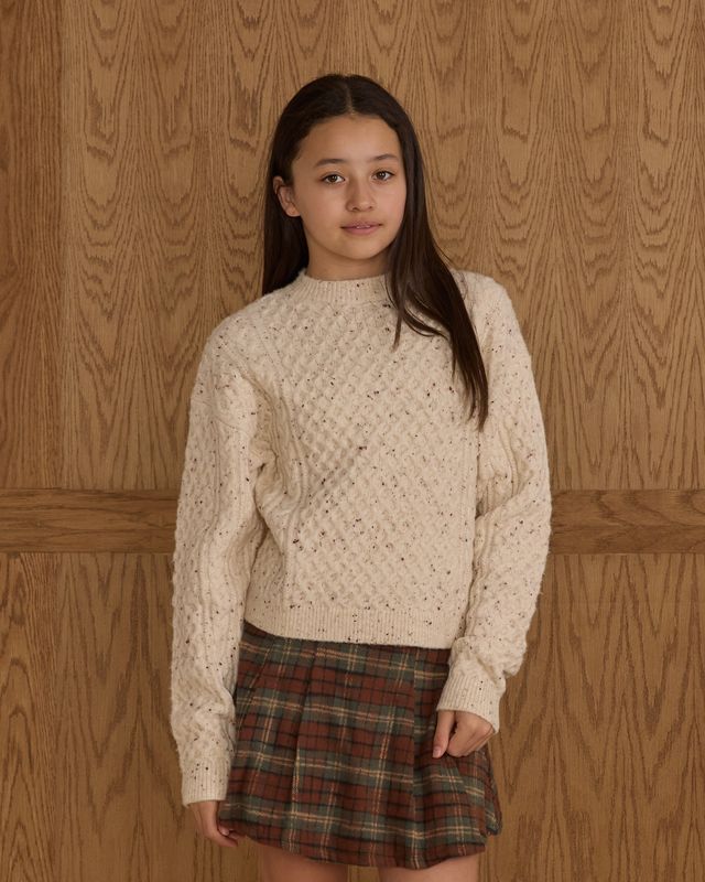 Young girl wearing a cream sweater and plaid skirt against a wooden paneled wall.