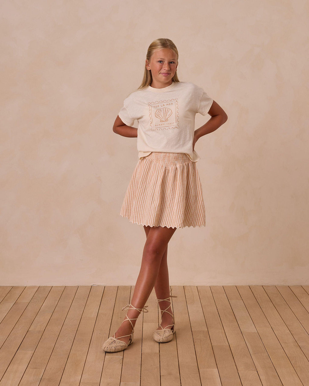 young girl modeling the boxy tee with seashell printed on the front of the top in a grapefruit color. she also has the grapefruit seersucker skirt on. 