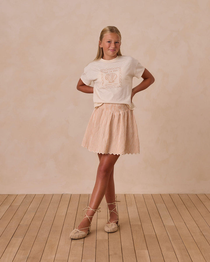 young girl modeling the boxy tee with seashell printed on the front of the top in a grapefruit color. she also has the grapefruit seersucker skirt on. 
