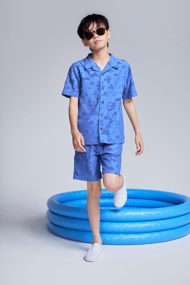 Child wearing a blue outfit standing next to a blue inflatable pool on a plain background