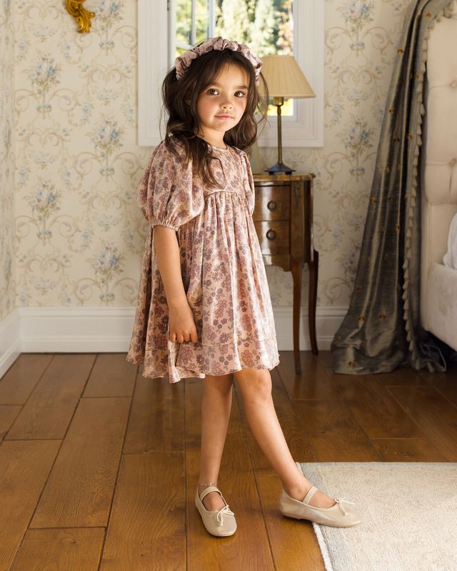 Young girl in a floral dress standing in a room with wooden floor and decorative wallpaper.