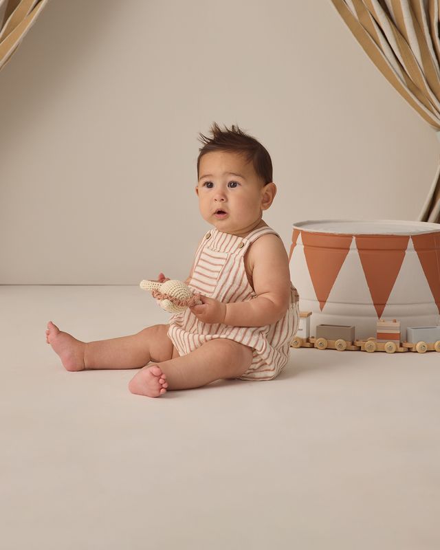 Baby in a striped romper sitting on a beige floor with a decorative container in the background.