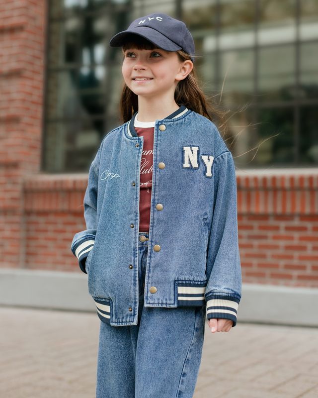Child wearing a blue 'NY' letterman jacket and cap outdoors.
