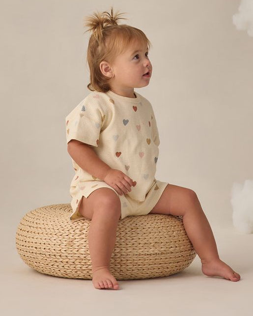 Child sitting on a woven stool wearing a light-colored outfit with heart patterns against a neutral background