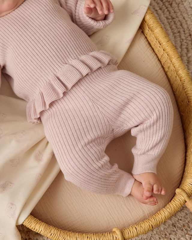 Baby in lilac ribbed outfit lying in a woven basket with beige blanket