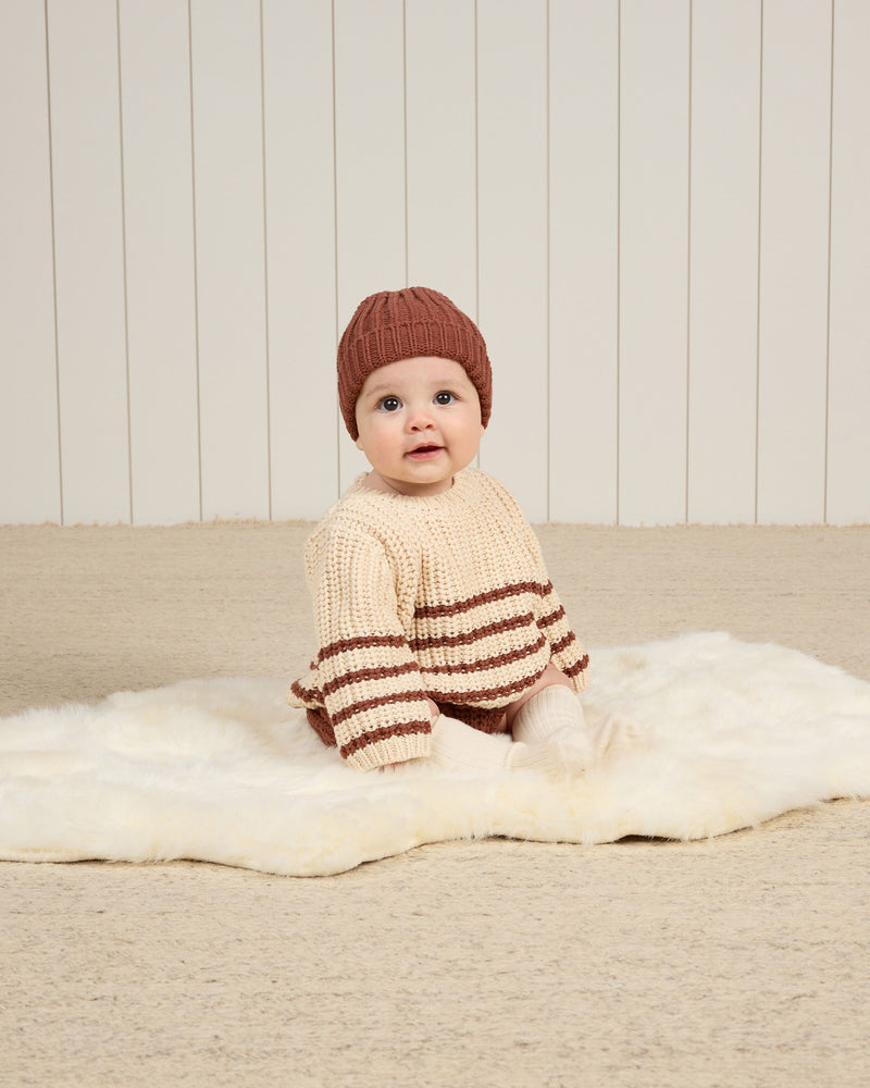 Baby modeling the chunky knit sweater with beige background and cranberry stripes on the bottom. 