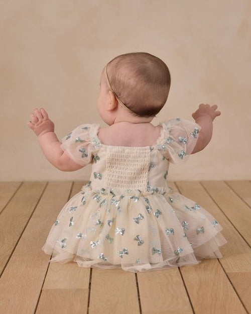 Baby in a floral dress standing on a wooden floor with a beige wall background