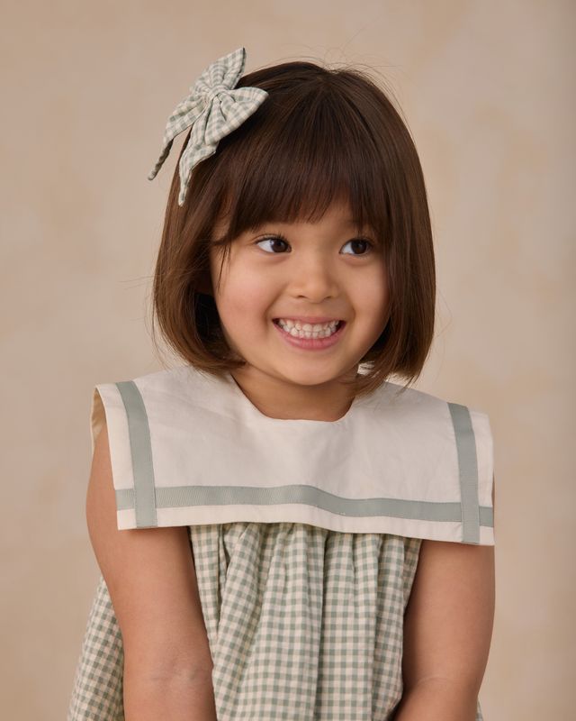 Young girl wearing a checkered dress with a bow in her hair against a beige background