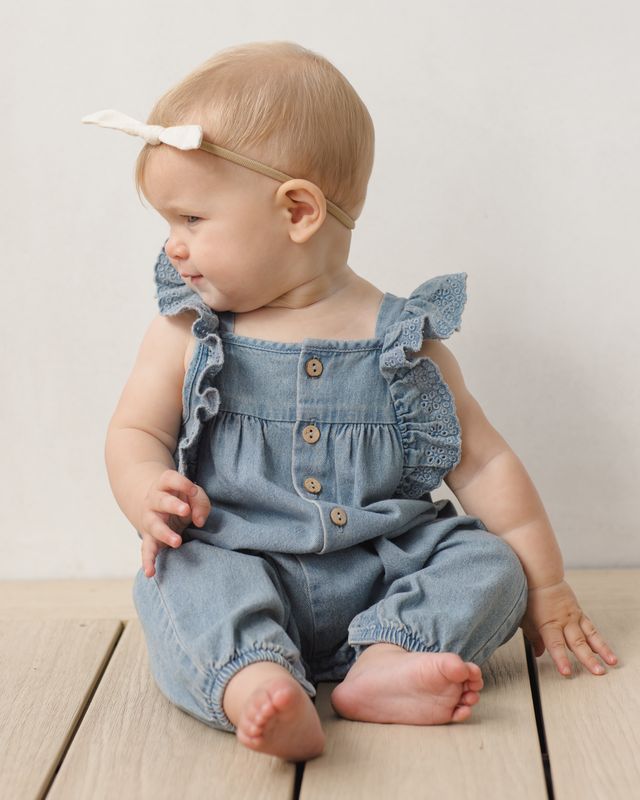 Baby wearing a denim romper with ruffled straps on a wooden floor.