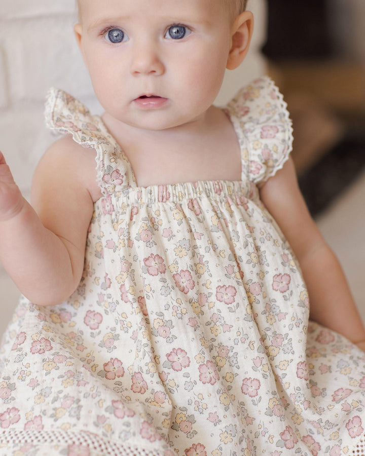 baby wearing the ruffle tank dress in a pastel floral print. the colors in the dress are pink, yellow and sage green. 